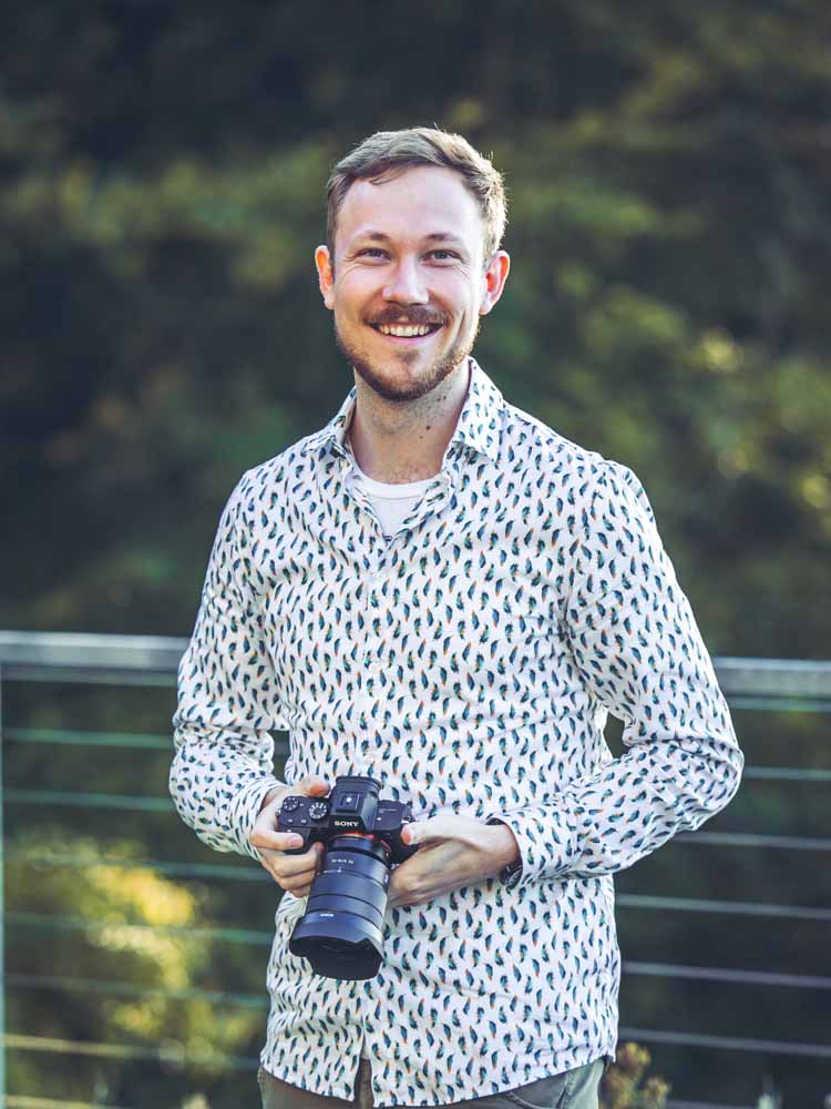 Lächelnder Fotograf Eric Bubacz hält eine Kamera und steht vor einem natürlichen grünen Hintergrund.