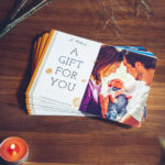 Elegante Geschenkgutscheine von Eric Bubacz Fotografie auf einem Holztisch, mit einem liebevollen Bild eines Paares, das ihr Neugeborenes betrachtet, neben sanfter Pflanzendekoration und einer warmen Kerzenflamme.