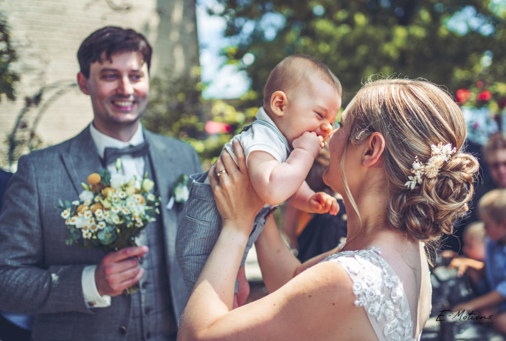 Eine fröhliche Braut in einem eleganten Hochzeitskleid küsst ihr lachendes Baby, während der Bräutigam neben ihnen steht, vor dem historischen Hintergrund der Godesburg in Bad Godesberg, festgehalten von Eric Bubacz Fotografie.